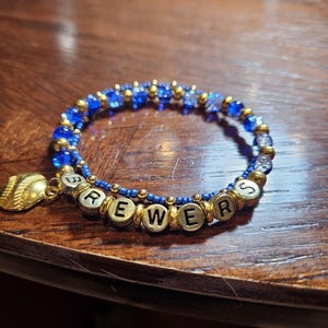 May include: A beaded bracelet featuring blue and clear glass beads, small gold-toned spacer beads, and silver-toned letter beads spelling out the word BREWERS. A gold-toned baseball charm is attached to the side of the circular bracelet.