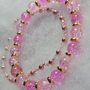 May include: Two pink beaded bracelets. The larger bracelet features cracked glass beads with gold-colored spacers. The smaller bracelet has small, round, rose-gold colored beads. The bracelets are on a white, textured surface.