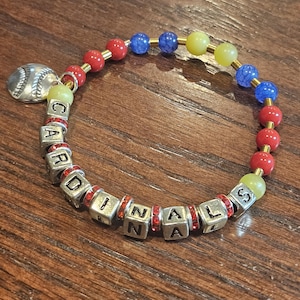 May include: A beaded bracelet with the word "CARDINALS" spelled out in silver letter beads. The bracelet features red, blue, and yellow beads, with a baseball charm.