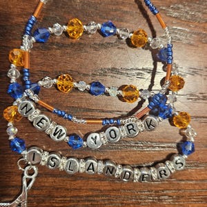May include: Three beaded bracelets featuring blue, orange, and clear crystal beads. One bracelet displays the text NEW YORK, another displays ISLANDERS, and a third features a silver-toned hockey stick charm. The beads are arranged in alternating patterns.