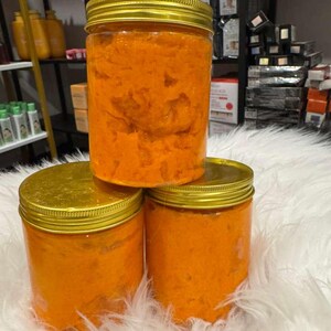 Op de afbeelding: Drie doorzichtige potten met gouden deksels gevuld met een oranje, getextureerde substantie. De potten zijn gestapeld op een witte, donzige ondergrond. De achtergrond toont planken met diverse producten.