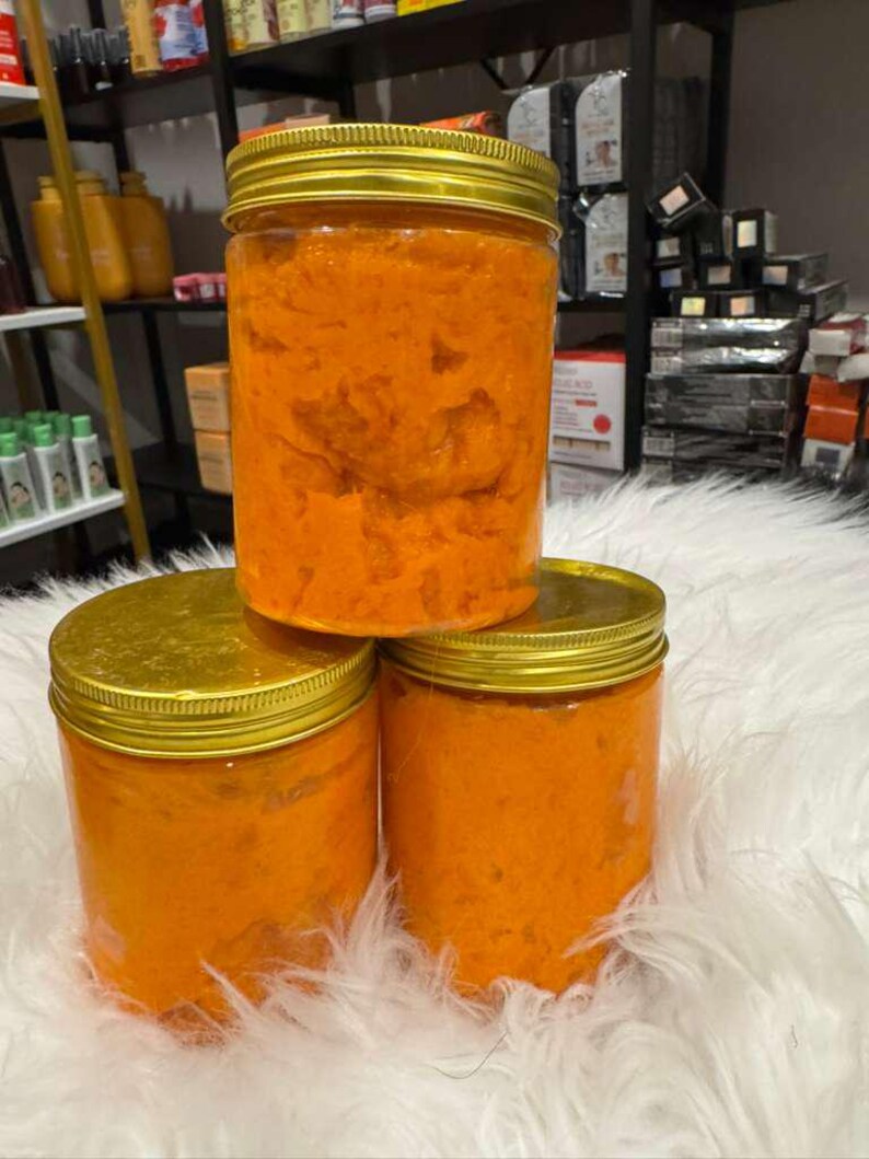 Op de afbeelding: Drie doorzichtige potten met gouden deksels gevuld met een oranje, getextureerde substantie. De potten zijn gestapeld op een witte, donzige ondergrond. De achtergrond toont planken met diverse producten.