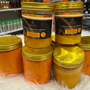 Op de afbeelding: Verschillende doorzichtige potten met gouden deksels gevuld met oranje en gele cr&egrave;mes. De potten hebben etiketten met zwarte en gouden tekst met de tekst "ADOJA SAVON ECLAIRCISSANT SUPREME". De potten staan op een wit oppervlak.