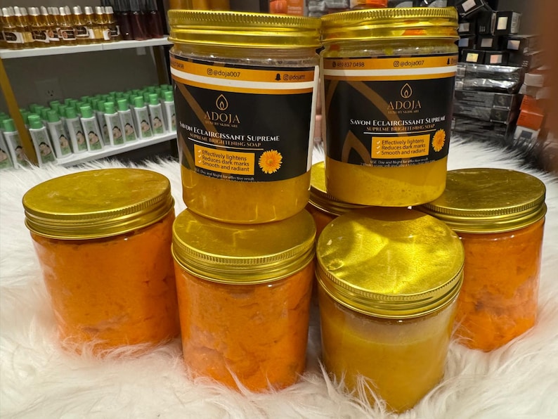 Op de afbeelding: Verschillende doorzichtige potten met gouden deksels gevuld met oranje en gele cr&egrave;mes. De potten hebben etiketten met zwarte en gouden tekst met de tekst "ADOJA SAVON ECLAIRCISSANT SUPREME". De potten staan op een wit oppervlak.