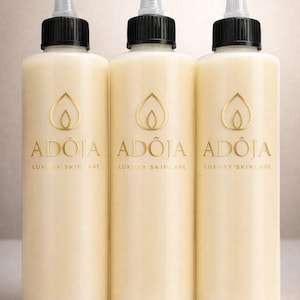 May include: Three cream-colored skincare bottles with black caps and clear dispensing tips. Each bottle has a gold logo and the text "AD&Ocirc;JA LUXURY SKINCARE". The bottles are cylindrical and appear to be filled with a creamy lotion.