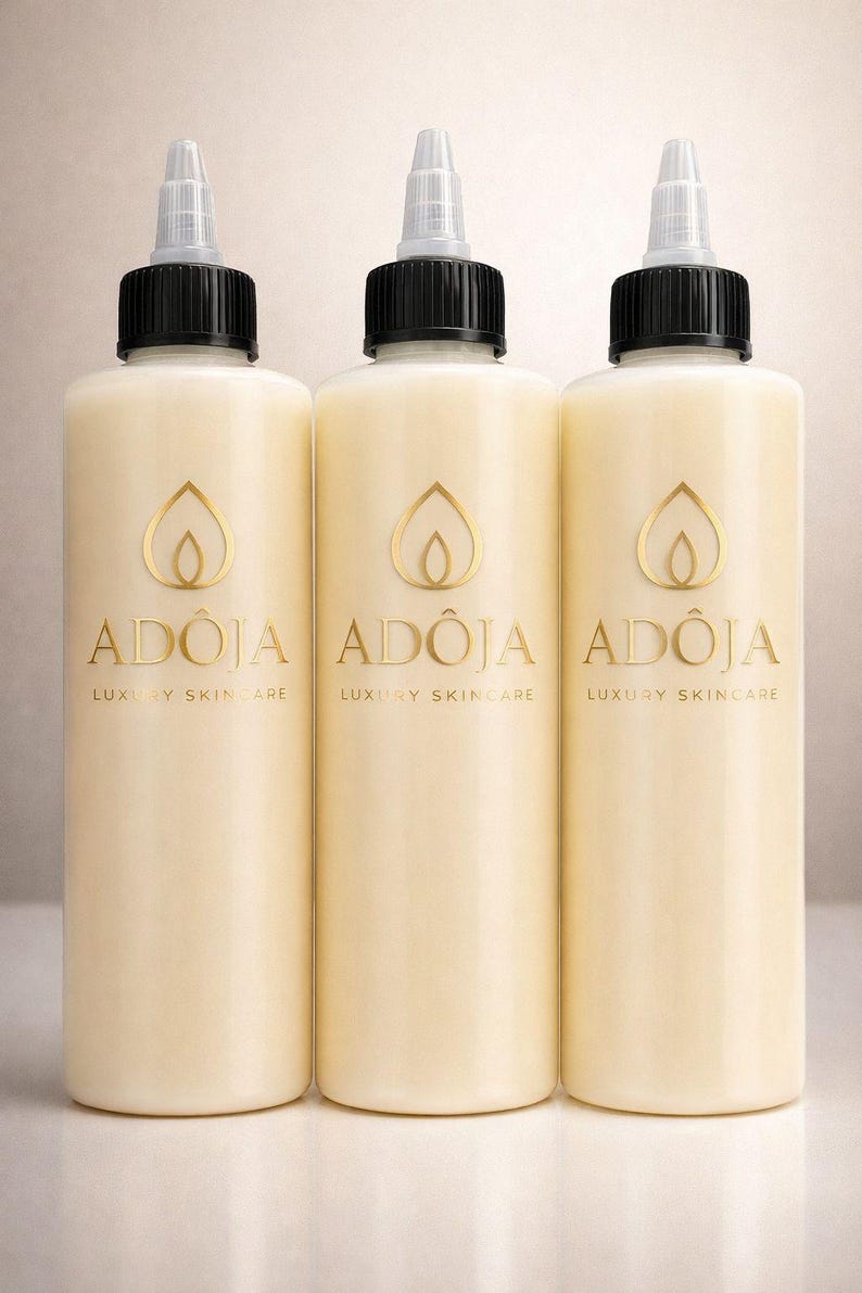 May include: Three cream-colored skincare bottles with black caps and clear dispensing tips. Each bottle has a gold logo and the text "AD&Ocirc;JA LUXURY SKINCARE". The bottles are cylindrical and appear to be filled with a creamy lotion.