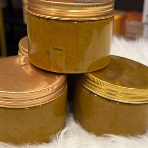 Op de afbeelding: Een stapel van vier doorzichtige potten gevuld met een dikke, geelbruine substantie. Elke pot heeft een goudkleurige deksel en een gouden rand. De potten zijn gestapeld op een witte, zachte ondergrond.