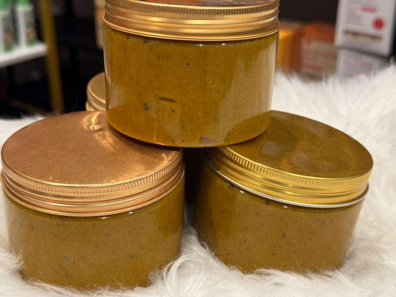 Op de afbeelding: Een stapel van vier doorzichtige potten gevuld met een dikke, geelbruine substantie. Elke pot heeft een goudkleurige deksel en een gouden rand. De potten zijn gestapeld op een witte, zachte ondergrond.
