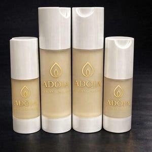 May include: Four skincare product bottles with white caps and a beige base. The bottles have a gold logo and the brand name "ADOIA LUXURY SKINCARE" printed on them. The background is black.