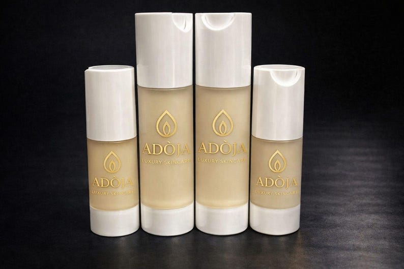 May include: Four skincare product bottles with white caps and a beige base. The bottles have a gold logo and the brand name "ADOIA LUXURY SKINCARE" printed on them. The background is black.