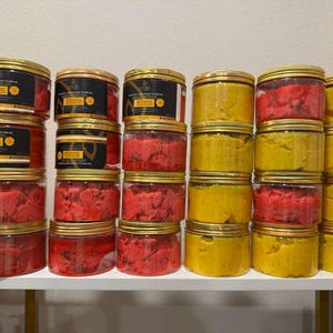 Op de afbeelding: Een display van transparante potten met gouden deksels, gevuld met rode en gele producten. Sommige potten hebben etiketten met zwarte en gouden tekst. De potten zijn gestapeld op een witte plank.