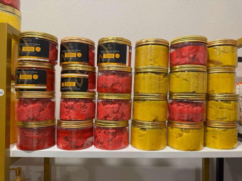 Op de afbeelding: Een display van transparante potten met gouden deksels, gevuld met rode en gele producten. Sommige potten hebben etiketten met zwarte en gouden tekst. De potten zijn gestapeld op een witte plank.