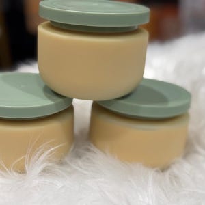 Op de afbeelding: Drie kleine, ronde containers met lichtgroene deksels gestapeld op een zacht, wit oppervlak. De containers zijn lichtbeige van kleur. De deksels zijn een gedempt groen. De containers zijn op elkaar gestapeld.