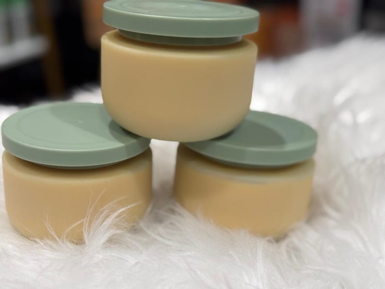 Op de afbeelding: Drie kleine, ronde containers met lichtgroene deksels gestapeld op een zacht, wit oppervlak. De containers zijn lichtbeige van kleur. De deksels zijn een gedempt groen. De containers zijn op elkaar gestapeld.