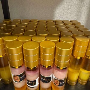 Op de afbeelding: Een plank vol met talrijke cilindrische flessen. De flessen hebben goudkleurige doppen en bodems, met een heldere body die een gelaagde vloeistof onthult. Sommige flessen hebben etiketten met een vrouwenafbeelding en tekst. De kleurstelling is goud, roze en helder.