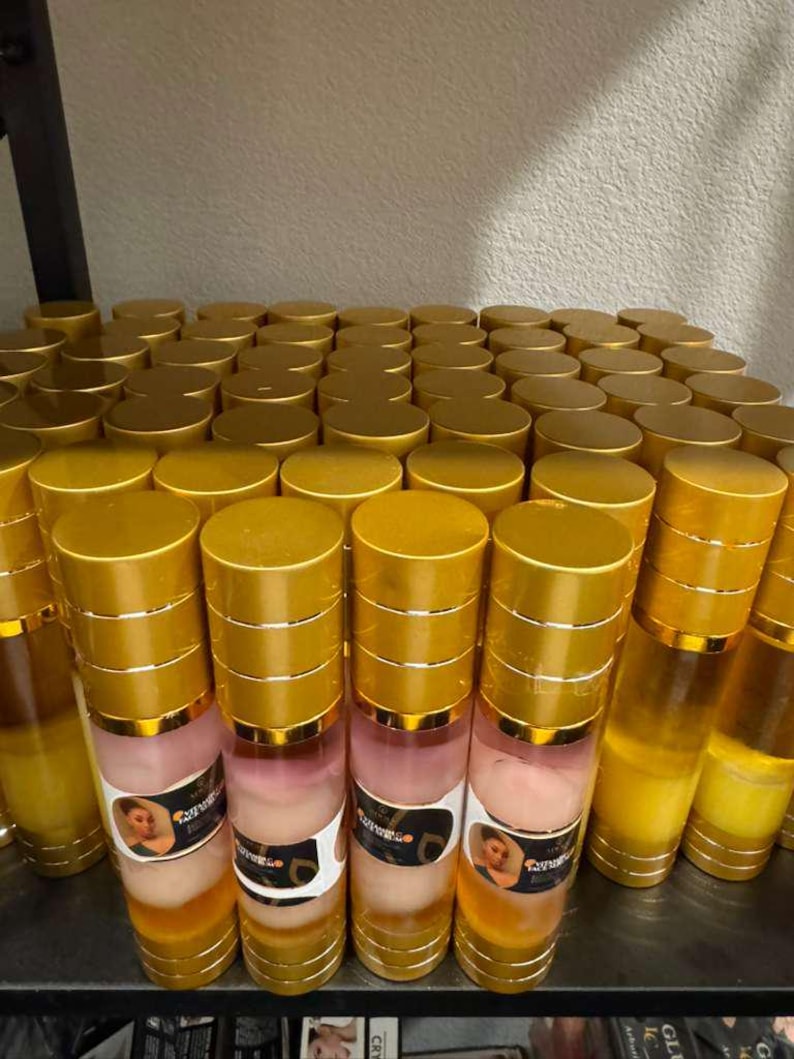 Op de afbeelding: Een plank vol met talrijke cilindrische flessen. De flessen hebben goudkleurige doppen en bodems, met een heldere body die een gelaagde vloeistof onthult. Sommige flessen hebben etiketten met een vrouwenafbeelding en tekst. De kleurstelling is goud, roze en helder.