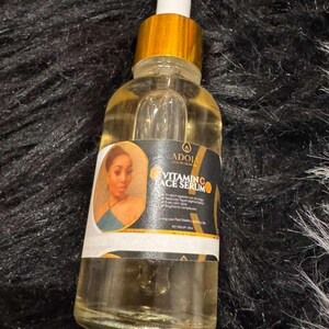 Op de afbeelding: Een heldere glazen fles met vitamine C gezichtsserum met een goudkleurige dop en druppelaar. Het etiket toont een vrouwenportret en de tekst "Vitamin C Face Serum". De fles bevat een gele vloeistof.