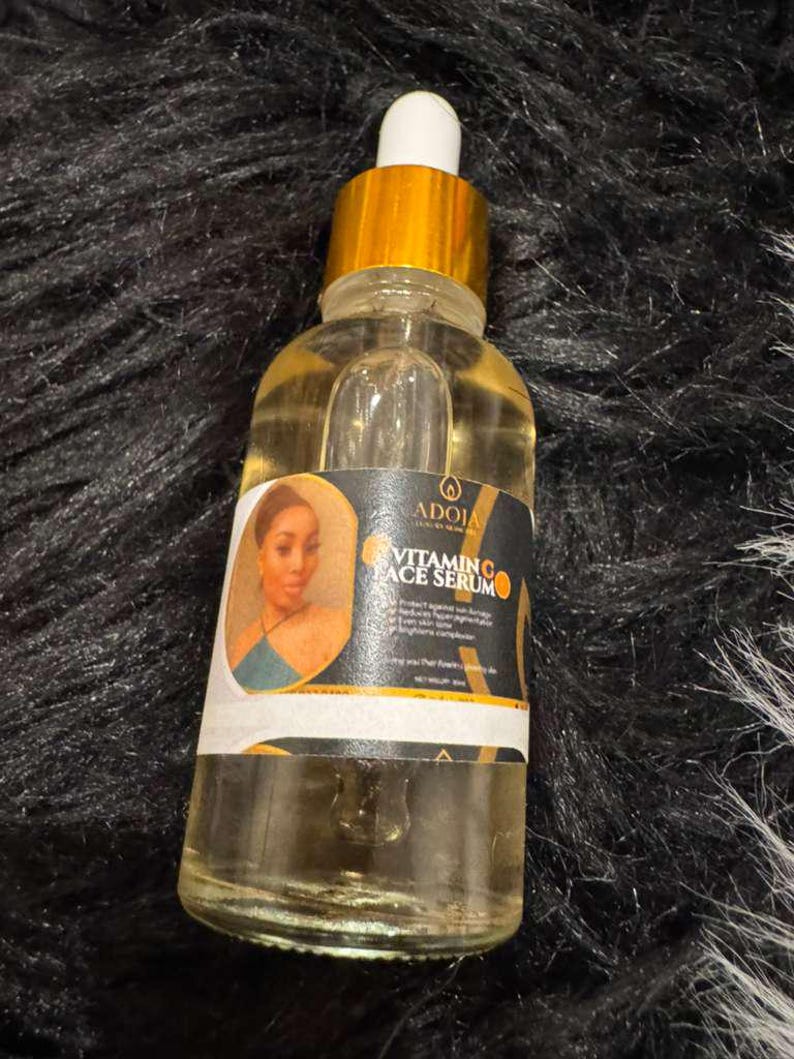Op de afbeelding: Een heldere glazen fles met vitamine C gezichtsserum met een goudkleurige dop en druppelaar. Het etiket toont een vrouwenportret en de tekst "Vitamin C Face Serum". De fles bevat een gele vloeistof.