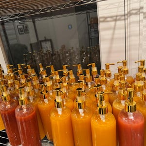 Op de afbeelding: Een verzameling heldere plastic flessen gevuld met kleurrijke vloeistoffen, gerangschikt op een metalen plank. Elke fles heeft een goudkleurige pompdosator. De vloeistoffen vari&euml;ren van rood tot oranje en geel.