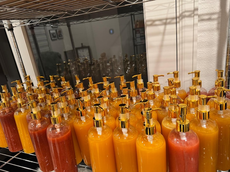 Op de afbeelding: Een verzameling heldere plastic flessen gevuld met kleurrijke vloeistoffen, gerangschikt op een metalen plank. Elke fles heeft een goudkleurige pompdosator. De vloeistoffen vari&euml;ren van rood tot oranje en geel.