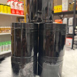 Op de afbeelding: Een stapel van zes zwarte, cilindrische containers met schroefdeksels. De containers zijn gemaakt van een glanzend materiaal en zijn gerangschikt op een witte, zachte ondergrond. Schappen met producten zijn zichtbaar op de achtergrond.