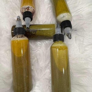 Op de afbeelding: Verschillende doorzichtige plastic flessen gevuld met een gele en groene vloeistof. De flessen hebben zwarte doppen met applicator tips. De flessen zijn gerangschikt op een witte, zachte ondergrond.