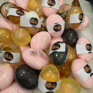 Op de afbeelding: Een verzameling verschillende zeepjes in eivorm. De zepen zijn in roze, geel en zwart, sommige met labels. De zepen zijn verpakt in doorzichtig plastic. De zepen liggen op een witte, zachte ondergrond.