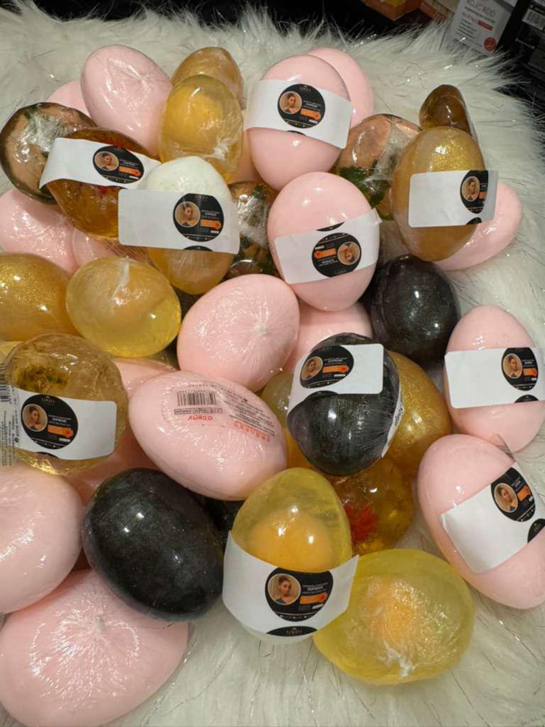Op de afbeelding: Een verzameling verschillende zeepjes in eivorm. De zepen zijn in roze, geel en zwart, sommige met labels. De zepen zijn verpakt in doorzichtig plastic. De zepen liggen op een witte, zachte ondergrond.