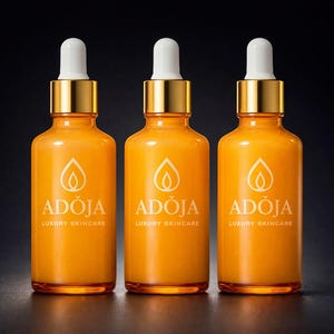 May include: Three orange glass skincare bottles with gold-colored caps and white droppers. Each bottle has the brand name "ADOJA" and "LUXURY SKINCARE" printed in white. The bottles are arranged in a row against a dark background.
