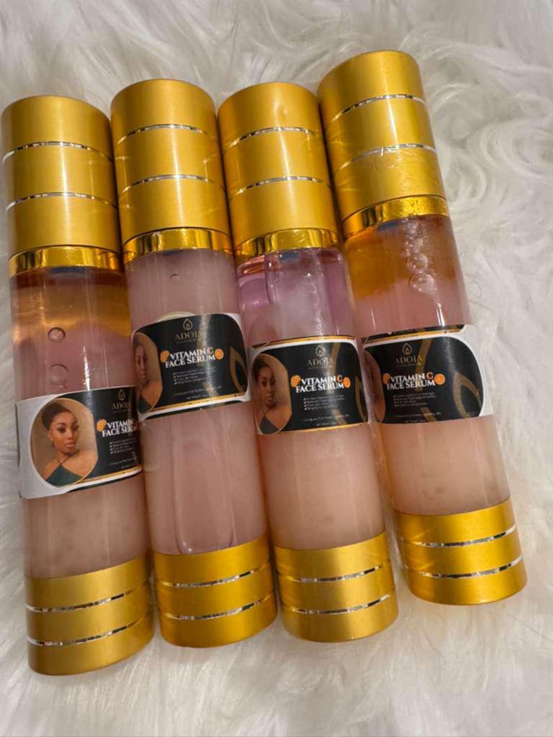Op de afbeelding: Vier doorzichtige flessen vitamine C gezichtsserum met goudkleurige doppen en bodems. Elke fles heeft een etiket met een portret van een vrouw en de tekst "Vitamin C Face Serum". Het serum binnenin is lichtroze van kleur.