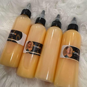 Op de afbeelding: Vier flessen Vital Face Serum met zwarte doppen en etiketten met een portret van een vrouw. De flessen zijn gevuld met een lichtoranje vloeistof en staan op een witte, zachte ondergrond.