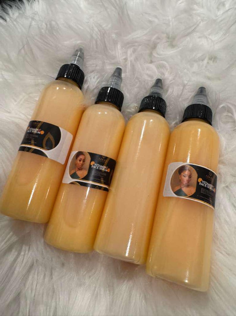 Op de afbeelding: Vier flessen Vital Face Serum met zwarte doppen en etiketten met een portret van een vrouw. De flessen zijn gevuld met een lichtoranje vloeistof en staan op een witte, zachte ondergrond.