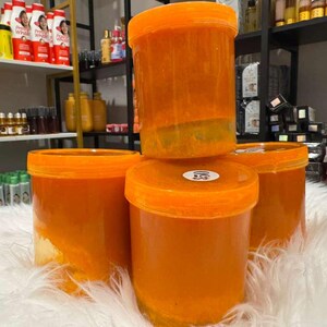 Op de afbeelding: Vier oranje plastic containers met oranje deksels zijn gestapeld op een witte, donzige ondergrond. De containers zijn gevuld met een oranje substantie. Schappen met diverse producten zijn zichtbaar op de achtergrond.