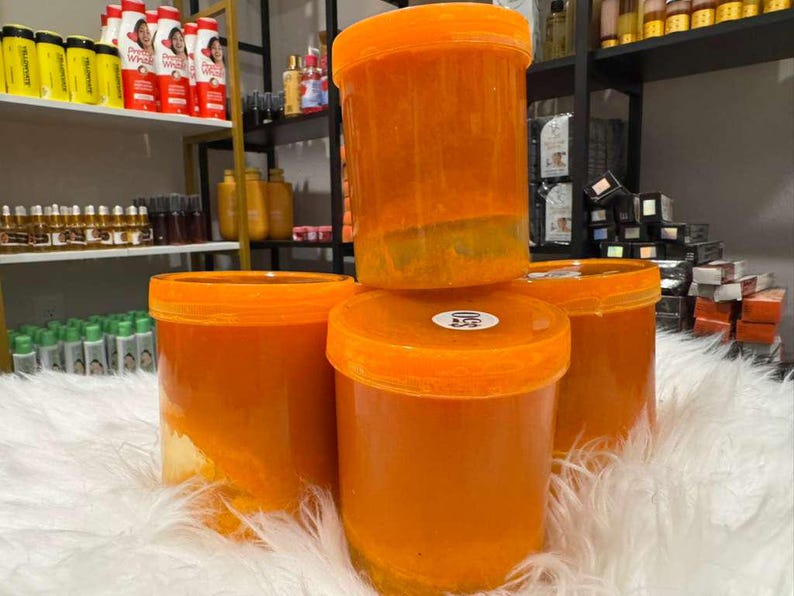Op de afbeelding: Vier oranje plastic containers met oranje deksels zijn gestapeld op een witte, donzige ondergrond. De containers zijn gevuld met een oranje substantie. Schappen met diverse producten zijn zichtbaar op de achtergrond.