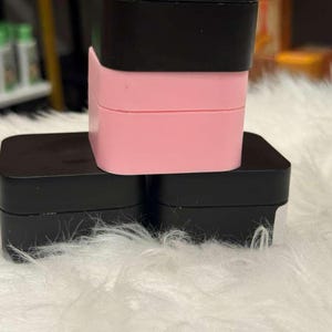 Op de afbeelding: Een stapel van drie vierkante containers. De onderste twee zijn zwart en de bovenste is roze, met een zwart deksel. De containers zijn gestapeld op een witte, donzige ondergrond.