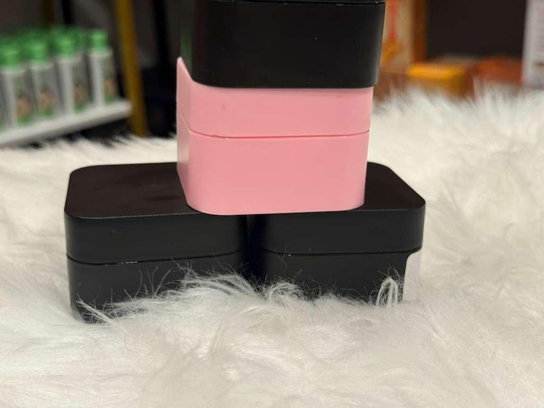 Op de afbeelding: Een stapel van drie vierkante containers. De onderste twee zijn zwart en de bovenste is roze, met een zwart deksel. De containers zijn gestapeld op een witte, donzige ondergrond.