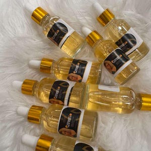 Op de afbeelding: Verschillende heldere glazen flessen gevuld met een gouden vloeistof, elk met een wit etiket en een gouden druppelaar. De etiketten lezen "Vitamin C Face Serum" en bevatten een afbeelding. De flessen zijn gerangschikt op een witte, zachte ondergrond.
