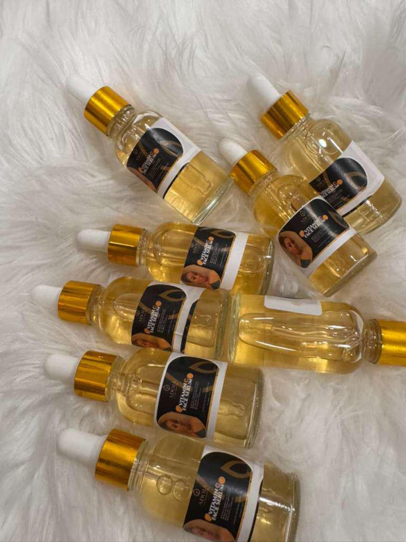 Op de afbeelding: Verschillende heldere glazen flessen gevuld met een gouden vloeistof, elk met een wit etiket en een gouden druppelaar. De etiketten lezen "Vitamin C Face Serum" en bevatten een afbeelding. De flessen zijn gerangschikt op een witte, zachte ondergrond.