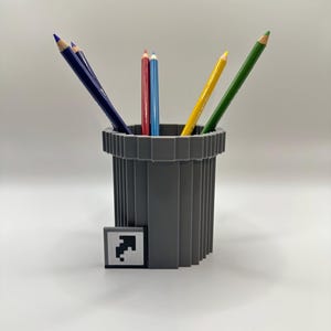 May include: A gray pencil holder with a ridged design, holding several colored pencils. The pencils are blue, red, yellow, and green. A small square tile with a black arrow is next to the holder. The pencils are arranged in a fan shape.