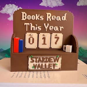 May include: A brown 3D-printed book counter with the text "Books Read This Year" and the number "017". It also features a "Stardew Valley" sign, two small book models, and a tiny mug. The background shows a purple and blue sky.