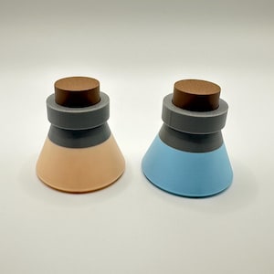 May include: Two decorative bottle stoppers. One is peach and gray, the other is blue and gray. Both have a wooden top and a gray band. The stoppers are cone-shaped and sit on a white surface.
