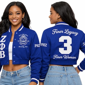 May include: A royal blue cropped varsity jacket featuring white striped ribbed trim. The front displays vertical white ZΦB lettering and a Zeta Phi Beta crest. The back features the number 3, the text Finer Legacy and Finer Woman, and PHZ on sleeves.