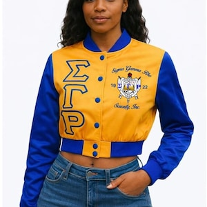 以下が含まれることがあります： ギリシャ文字のΣΓΡと「Sigma Gamma Rho」と「Sorority, Inc.」の文字が入った、黄色と青色のサテンのクロップドジャケット。 ジャケットは青い袖とトリム、青いスナップ留めが付いています。 画像の人物はブルージーンズを着用しています。