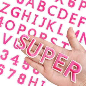 Puede incluir: Letras y números bordados en rosa sobre un fondo blanco. La palabra "SUPER" está escrita con letras individuales. Las letras y los números están en una fuente sans-serif.