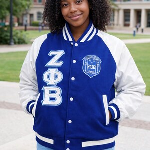 May include: A blue and white letterman jacket with the Greek letters ZΦB and a crest. The jacket has white sleeves, a blue body, and white trim. The person is wearing blue jeans.