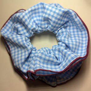 May include: A blue and white gingham scrunchie with a red trim. The fabric is gathered around an elastic band, creating a ruffled effect. This hair accessory is perfect for adding a touch of style to any hairstyle.