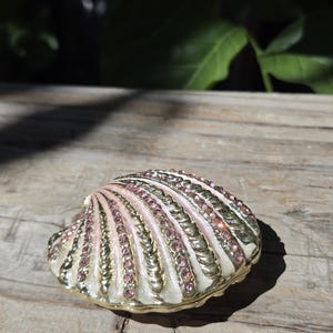 May include: A decorative trinket box shaped like a seashell, featuring a gold-tone metal finish with alternating ridges of pale pink enamel and rows of small, sparkling pink rhinestones embedded along the curved surface of the shell.