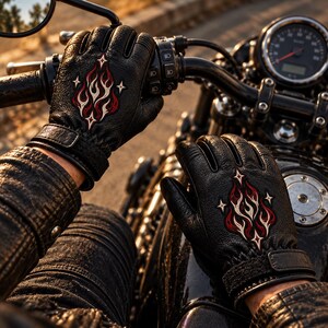 May include: Black leather motorcycle gloves featuring an embroidered design of red and white flames accented with four-pointed stars on the back of the hand. The gloves include a wrist strap with a hook-and-loop closure and textured leather detailing.