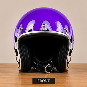 Puede incluir: Un casco de moto abierto de color morado con un diseño de pintura que gotea en tonos morados más claros y un patrón de cuadros blancos y negros en los laterales. El casco incluye un borde plateado, tres broches metálicos y una correa negra.