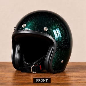 Può includere: Un casco da moto aperto con finitura glitter verde scuro. Il casco presenta un bordo nero, tre bottoni a pressione in metallo color argento sulla fronte e un sottogola nero con chiusura a doppio anello a D. La parola FRONT è stampata sotto.